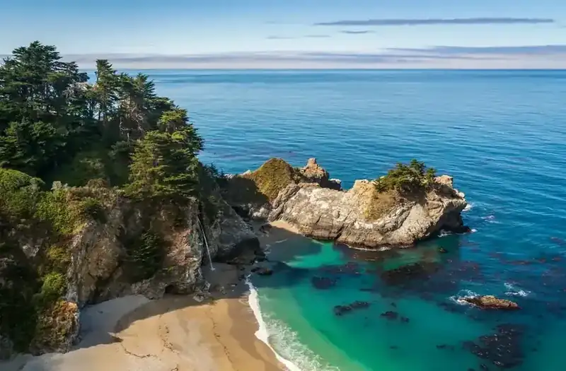 A waterfall dropping onto the sand of a turquoise cove on the Big Sur coast